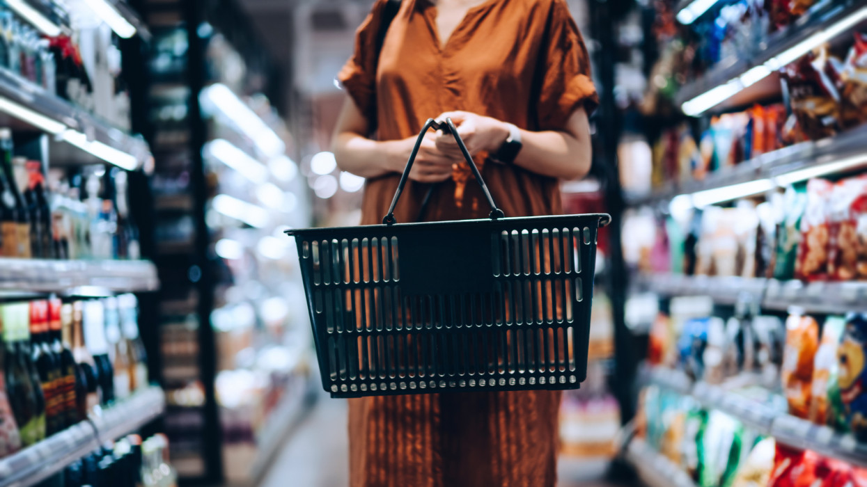 Cropped shot of young woman carrying a shopping basket, standing along the product aisle, grocery shopping for daily necessities in supermarket