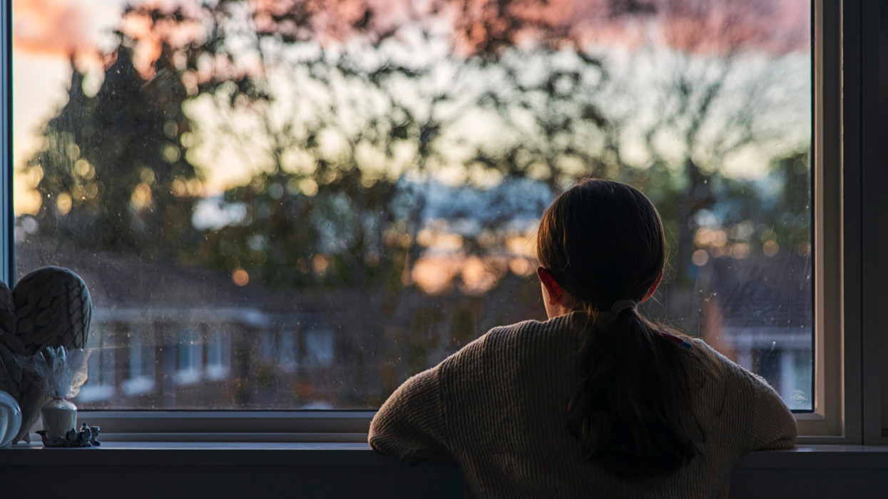 A young girl, with a ponytail, looks out of her bedroom window as the sun is setting. The view through the window is blurry but shows a residential area.