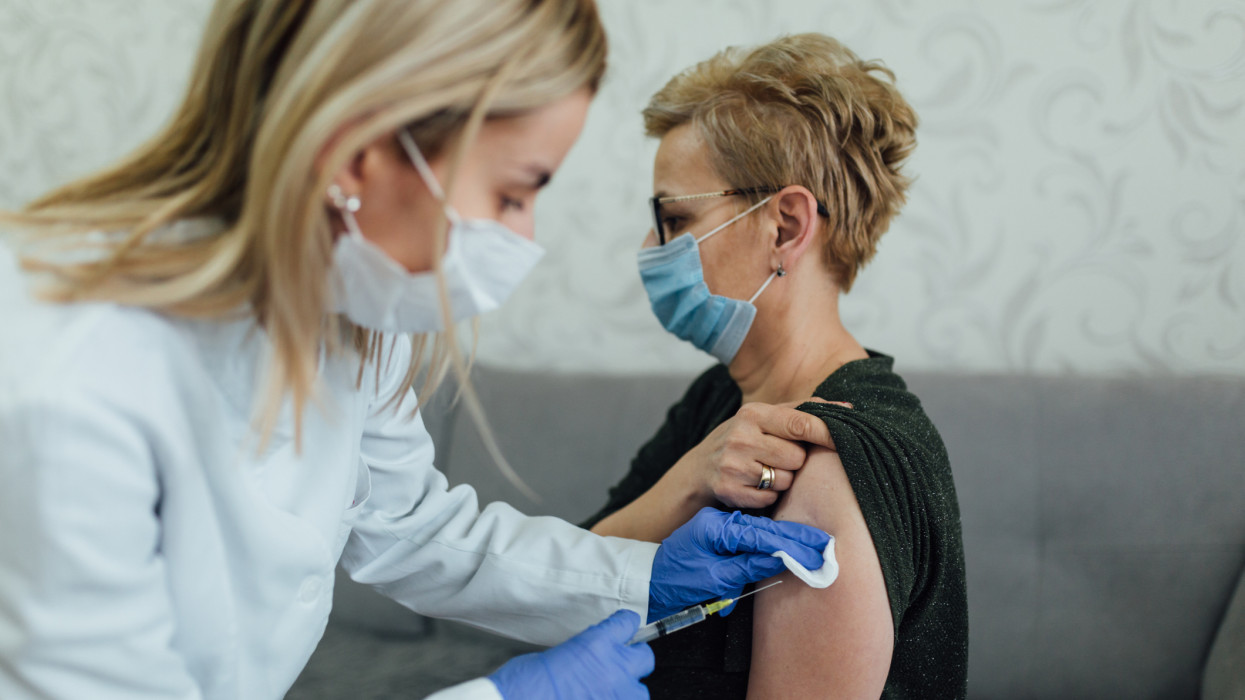 Unrecognizable senior Caucasian woman wearing a protective face mask and taking a vaccine in her left arm, the vaccine being administered by a female doctor wearing a protective face mask
