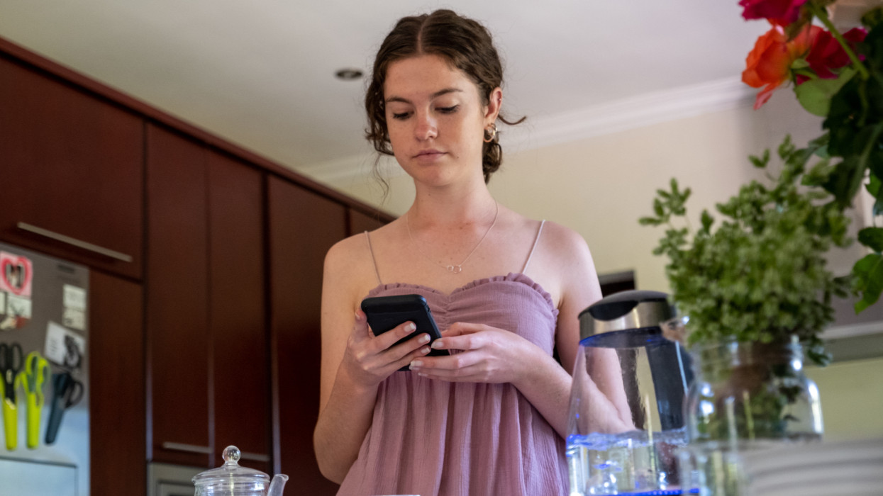 Young woman making herself some tea using loose tea. She uses her mobile phone as she waits for the tea to brew. Millennial generation enjoying a cup of tea