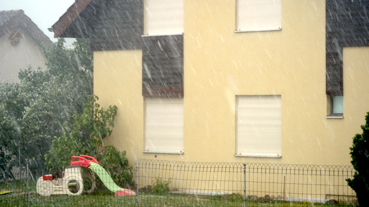 Heavy hailstorm beating against the facade of a house. There are hailstones on the lawn and the visibility is strongly reduced due to the dense rain and strong wind.