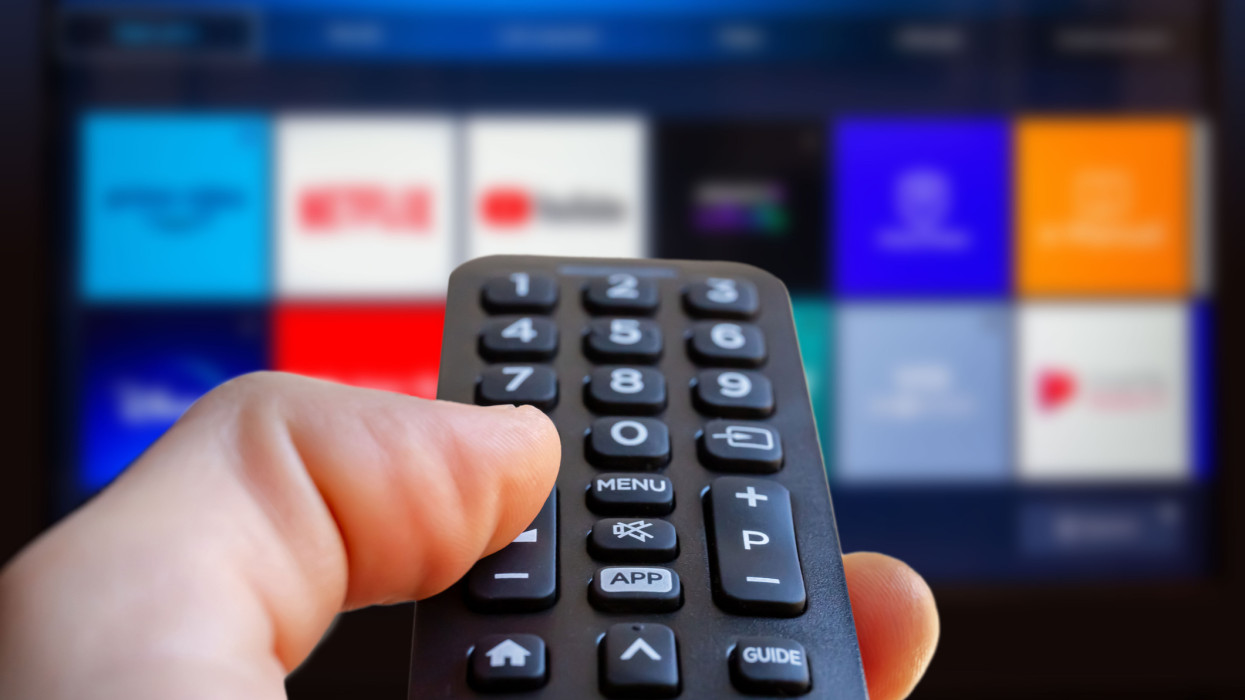 A man is holding a remote control of a smart TV in his hand. In the background you can see the television screen with streaming entertainment apps for video on demand