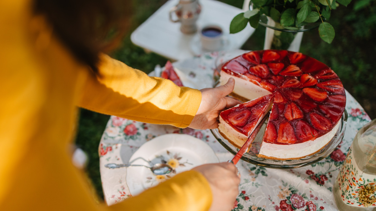 A young adult woman cutting a slice of a strawberry cheesecake on the well-decorated garden table with the beautiful floral tablecloth on