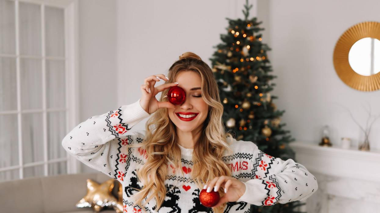 Portrait of a young cheerful woman with red lips and curly hair in a knitted sweater laughing and holding a red Christmas ball