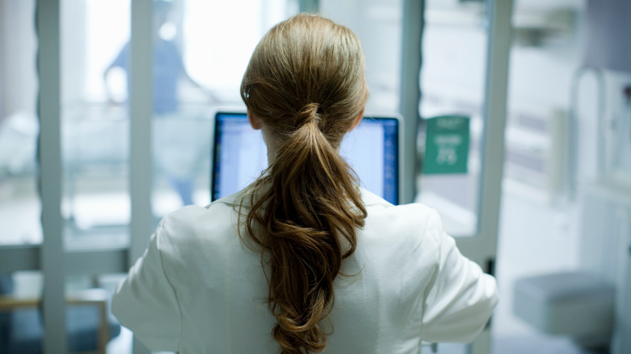 Woman doctor with long red hair using laptop in hospital, photographed from behind