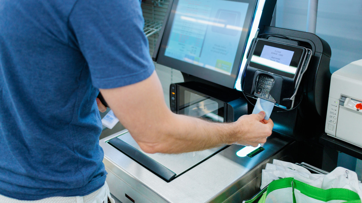Close-up of unrecognizable white man using credit card to purchase groceries at self-checkout kiosk in supermarket