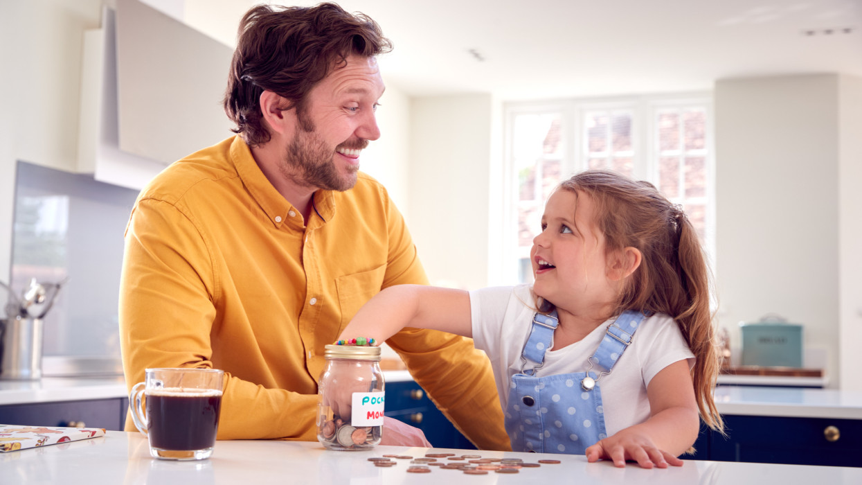Father And Daughter Counting Pocket Money In Jar On Kitchen Counter