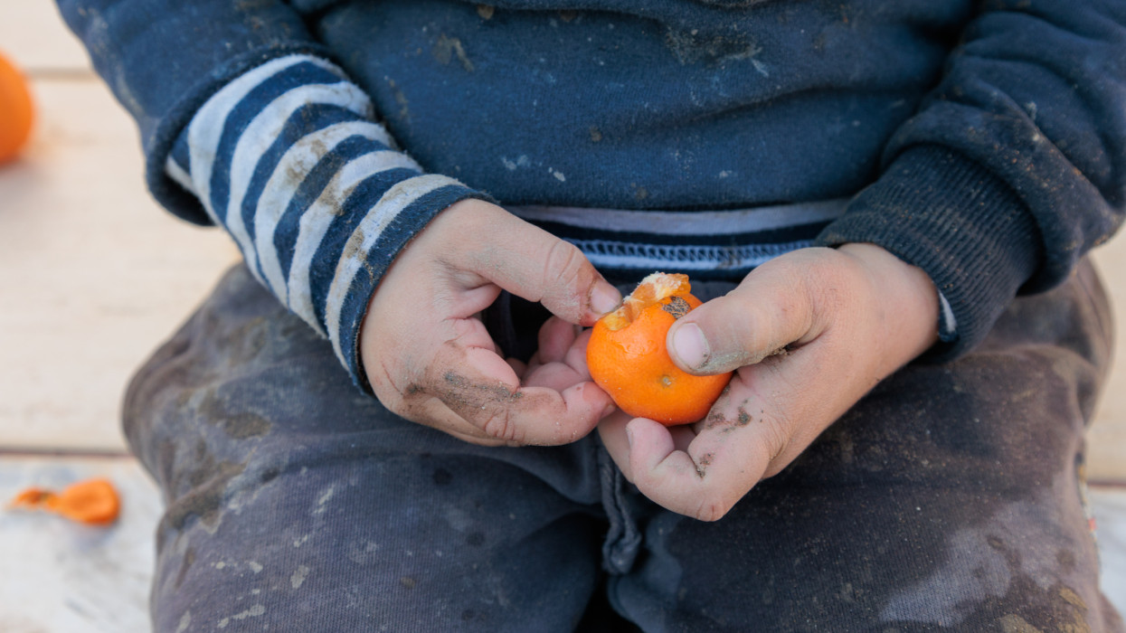 Preschool homeless boy covered in mud is holding tangerine for eating