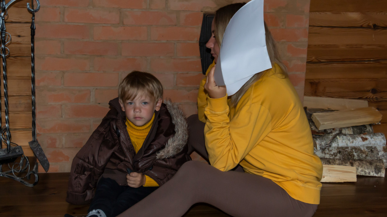 A woman with a small child dressed in warm clothes with bills in their hands and a mobile phone sit near a wood-burning, brick stove with birch firewood on the floor, in the interior of a wooden and environmentally friendly house in the winter season. Concept of ecology and energy and gas crisis.