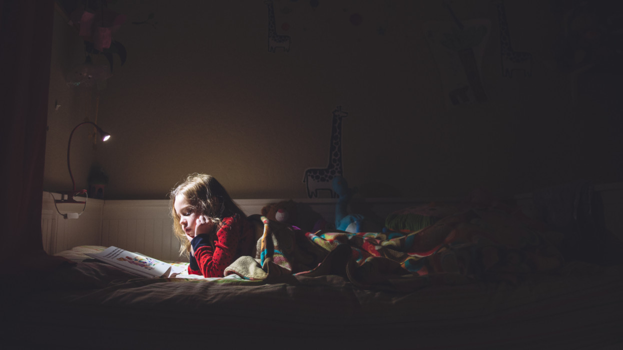 A six year old girl is reading in her bed at night.  The room is dark and she and her book are illuminated by a reading lamp.