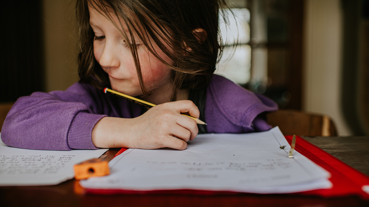 A little girl sits at a table. She looks as though she is putting in a lot of effort as she holds a pencil and attempts to problem solve. Space for copy.