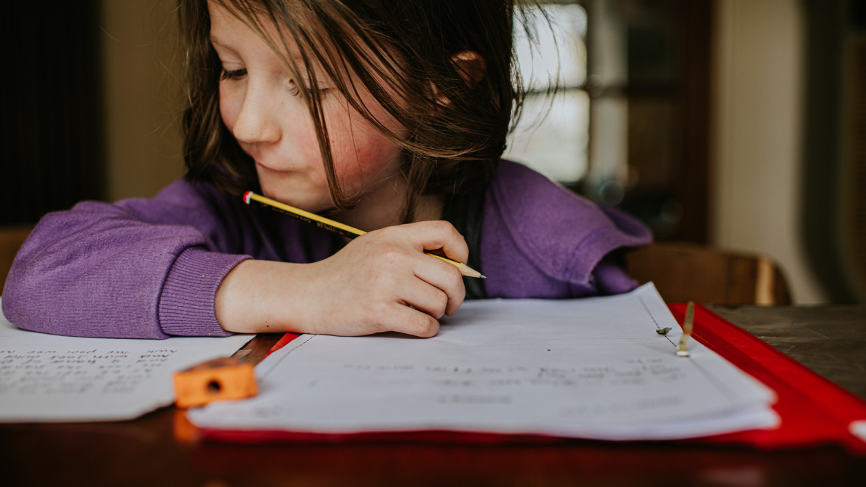 A little girl sits at a table. She looks as though she is putting in a lot of effort as she holds a pencil and attempts to problem solve. Space for copy.