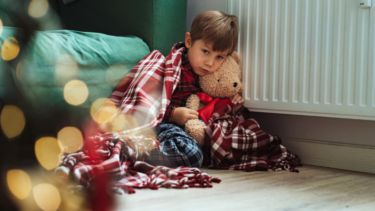 cute little boy wrapped id plaid falling asleep by heater hugging teddy bear in santa hat. Christmas time. Cold at home, Energy crisis. Fuel poverty