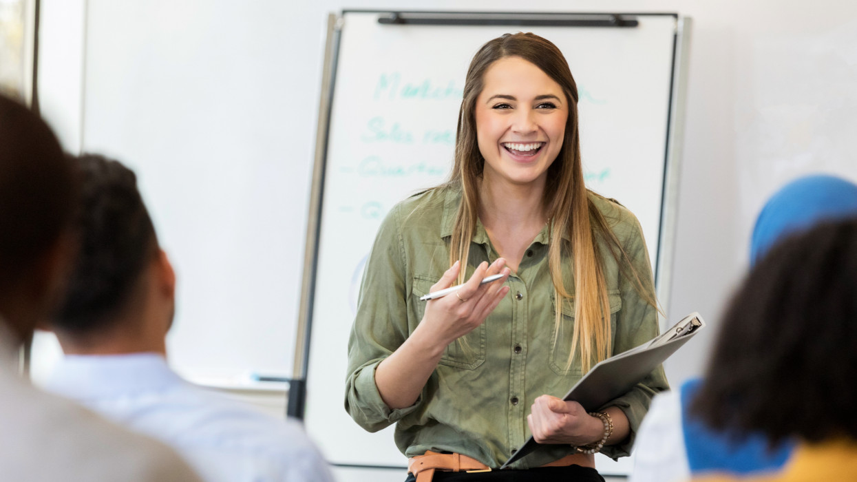 A smiling young female creative professional sits on a stool before a group of employees.  She gestures as she speaks to a new employee training class.