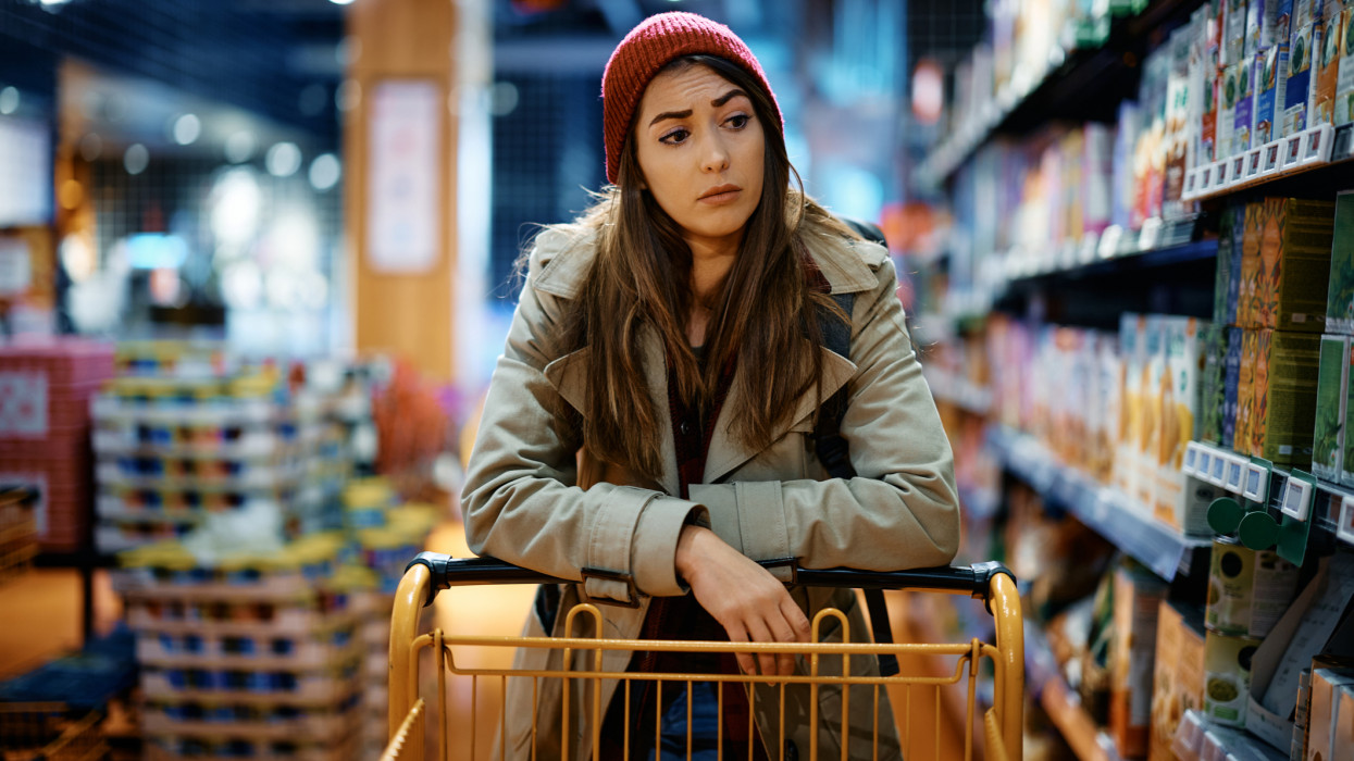 Young sad woman with empty shopping cart among produce aisle at supermarket.