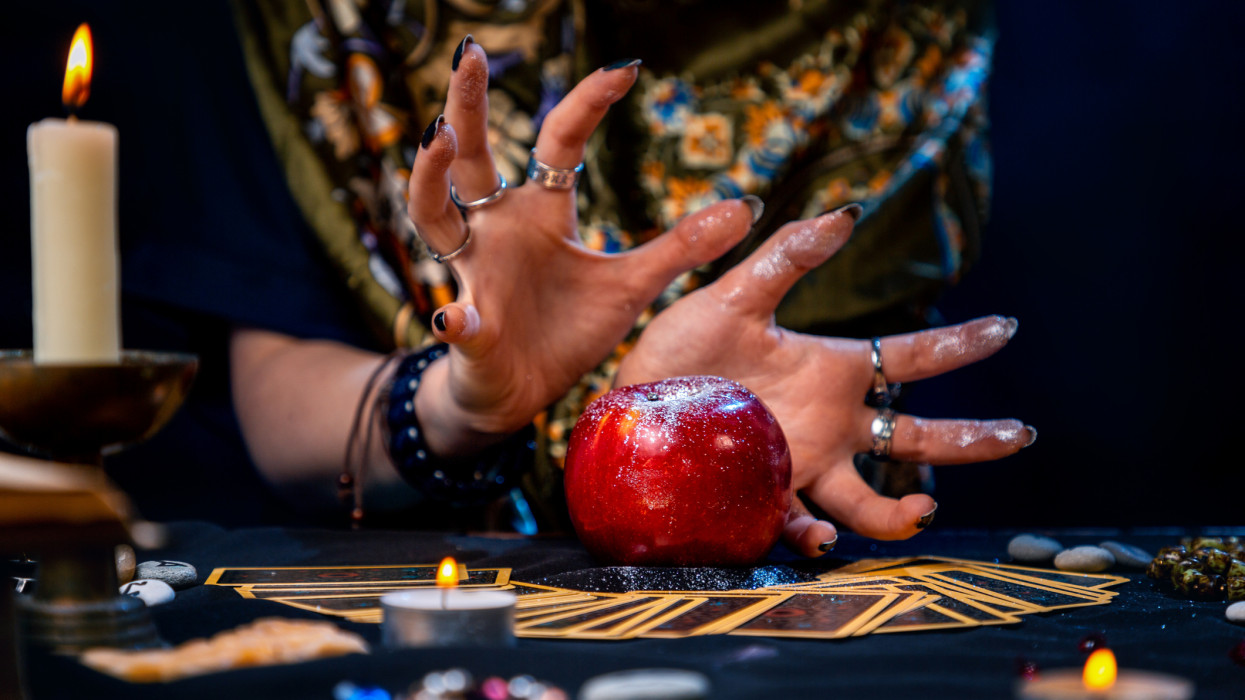 A fortune teller conjures a red apple lying on the Tarot cards. On the table is a lighted candle and amulets. Close up. The concept of divination, astrology and esotericism.