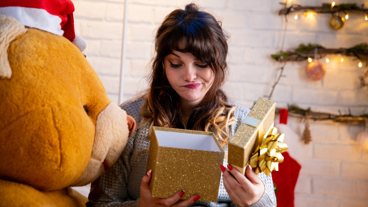Stylish happy girl opening Christmas gift box sitting on a giant bear. Girl opens Christmas gift. Woman and a giant teddy bear home alone for Christmas.
