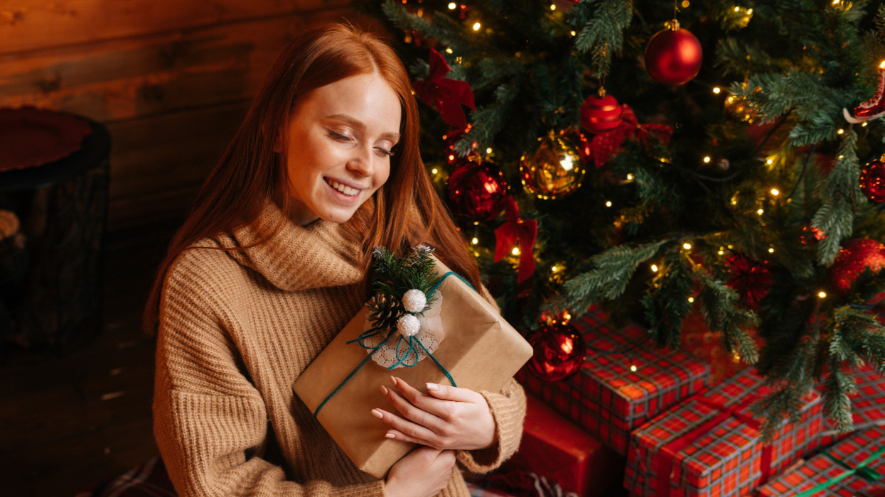 Close-up of smiling red-haired young woman with closed eyes holding gift box in hand received for Christmas sitting on floor background of xmas tree. Charming lady wearing sweater with holiday box.