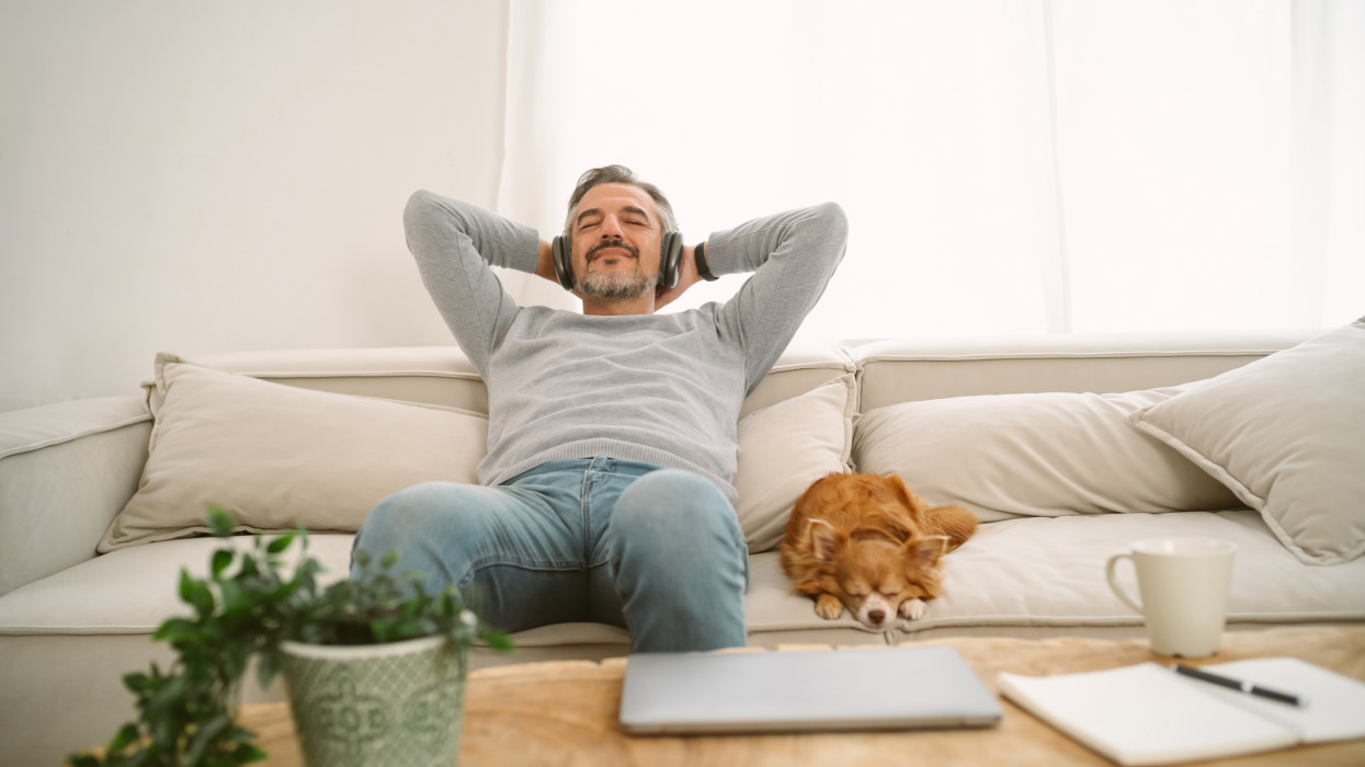 Calm Middle age Caucasian man sitting on sofa listening to music enjoying meditation for sleep and peaceful mind in wireless headphones, leaning back with his lovely chihuahua dog sit besides.