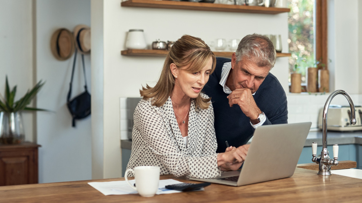 A senior couple planning their finance and paying bills while using a laptop at home. A mature man and woman going through paperwork and working online with a computer
