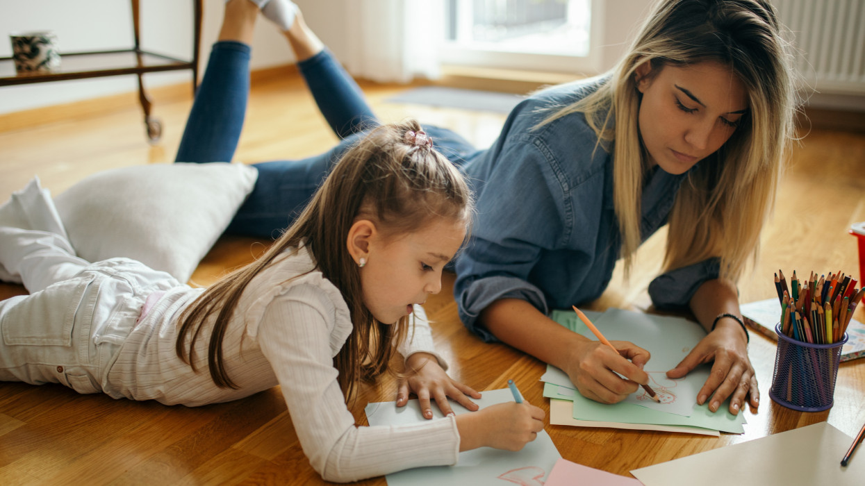 Young woman and little girl are playing and and drawing and coloring together in their living room while lying on floor. Woman is girls aunt and nanny. They are beautiful and playful