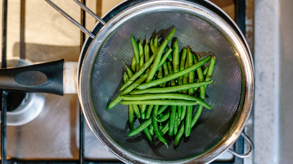 Close up of steamed green runner beans in a sieve on a kitchen stove top.