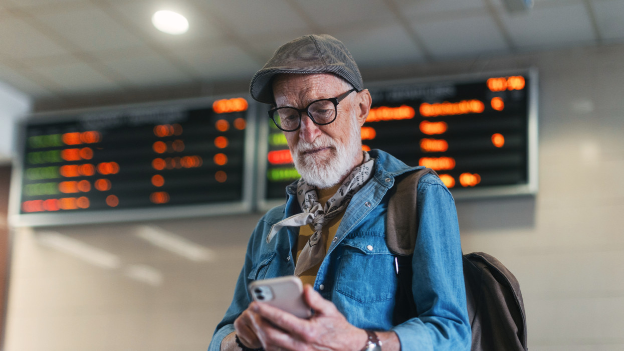 Stylish elderly man standing in train station under arrival and departure board. Senior man checking train delay, platform number, departure, arrival time on smartphone.