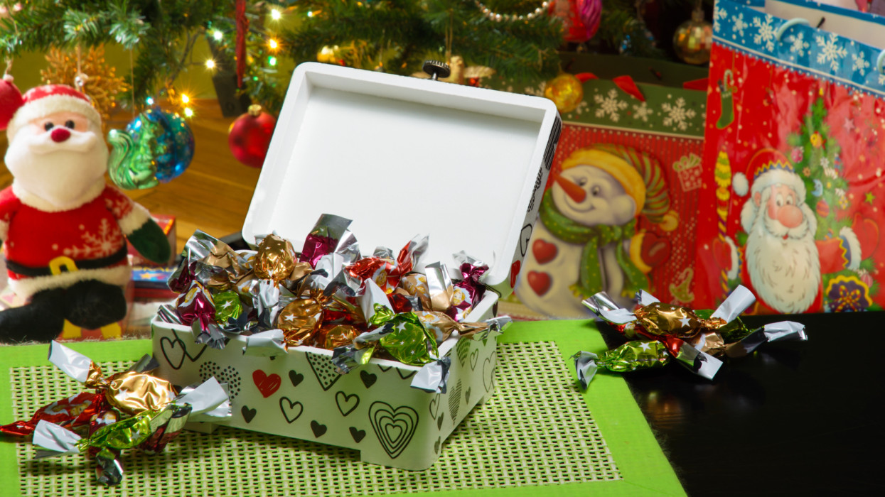 Decoupage box full of traditional Hungarian parlour candies. Beautifully decorated Christmas tree, gift bags under it, and a Santa Claus puppet are in a blurry background. Selective focus.