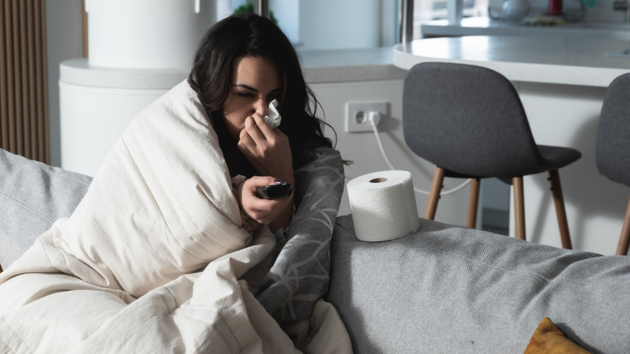 Young sick stressed woman lying at home covered with blanket blowing her nose. Ill female have common cold and flue with runny nose allergy reaction wipe it with paper tissue.
