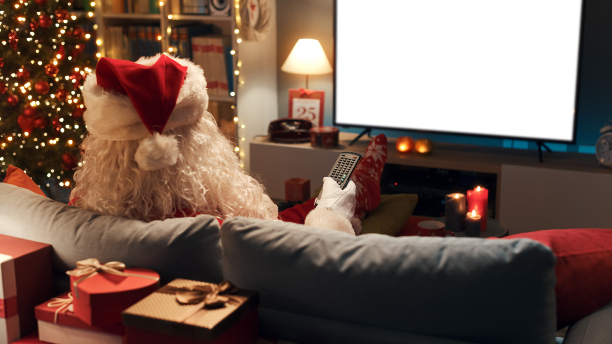 Santa Claus sitting on the sofa at home and watching tv, blank television screen with copy space