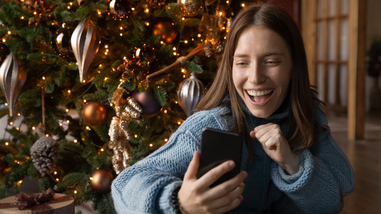 Overjoyed young caucasian woman looking at cellphone screen, feeling excited of winning New Year online lottery. Emotional millennial female blogger recording video on smartphone near Christmas tree.
