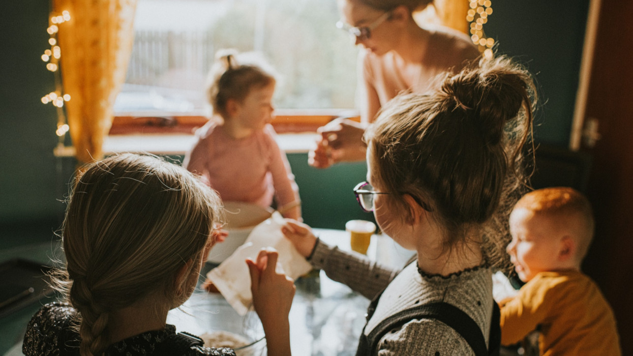 A mum of four kids sits at a table in a simple kitchen. She bakes with them, helping them to add ingredients and mix. The children are engaged in the activity. Scene is calm and loving. Two eldest girls in the foreground looking towards the rest of the family.