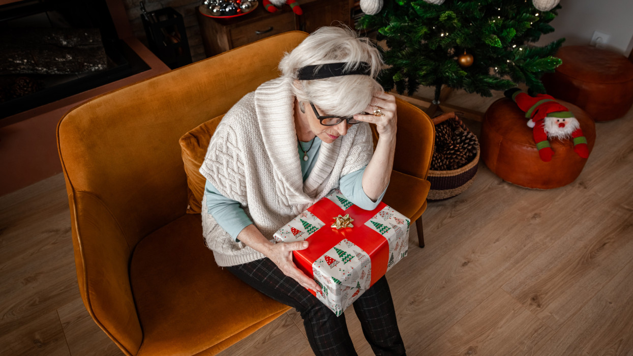 Top angle of Sad and bored senior woman, sitting alone in the living room with a wrapped gift. Sadness for Self isolation during Coronavirus pandemic for Christmas. Covid quarantine, lockdown.