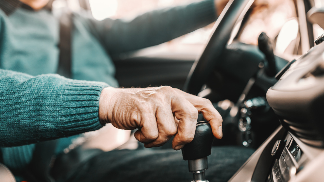 Close up of senior man holding one hand on gearshift and other on steering wheel while sitting in his car. Selective focus on hand.