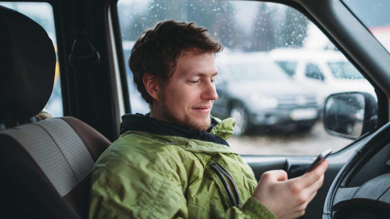 portrait of young smiling man in the green jacket holding smartphone in the car