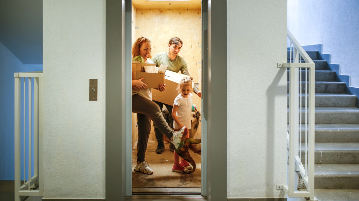 Family with one child moves with boxes with dog and toy dinosaur on elevator to new apartment
