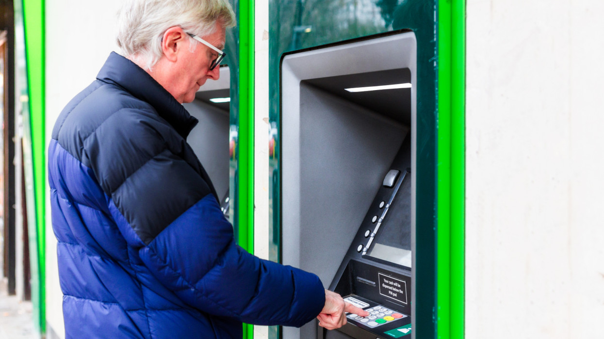 Color image depicting a senior man using an ATM machine on a city street.