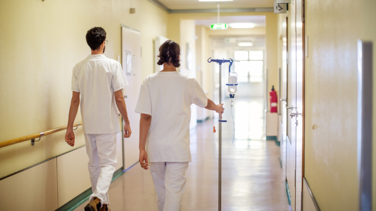 Rear view of two nurses walking through hospital hallway with saline stand. Medical staff in uniform walking through clinic corridor.