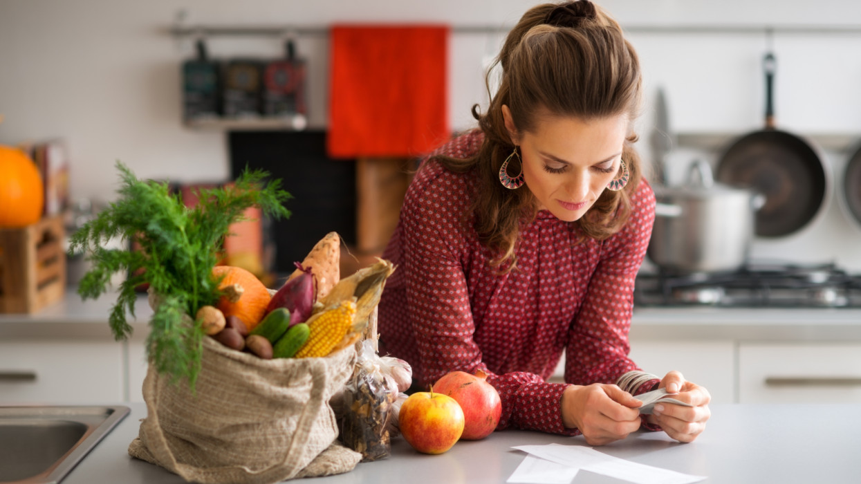 An elegant woman is reading the shopping lists on her kitchen counter. Next to her on the kitchen counter, a burlap sac holds a wide variety of fall fruits and vegetables.