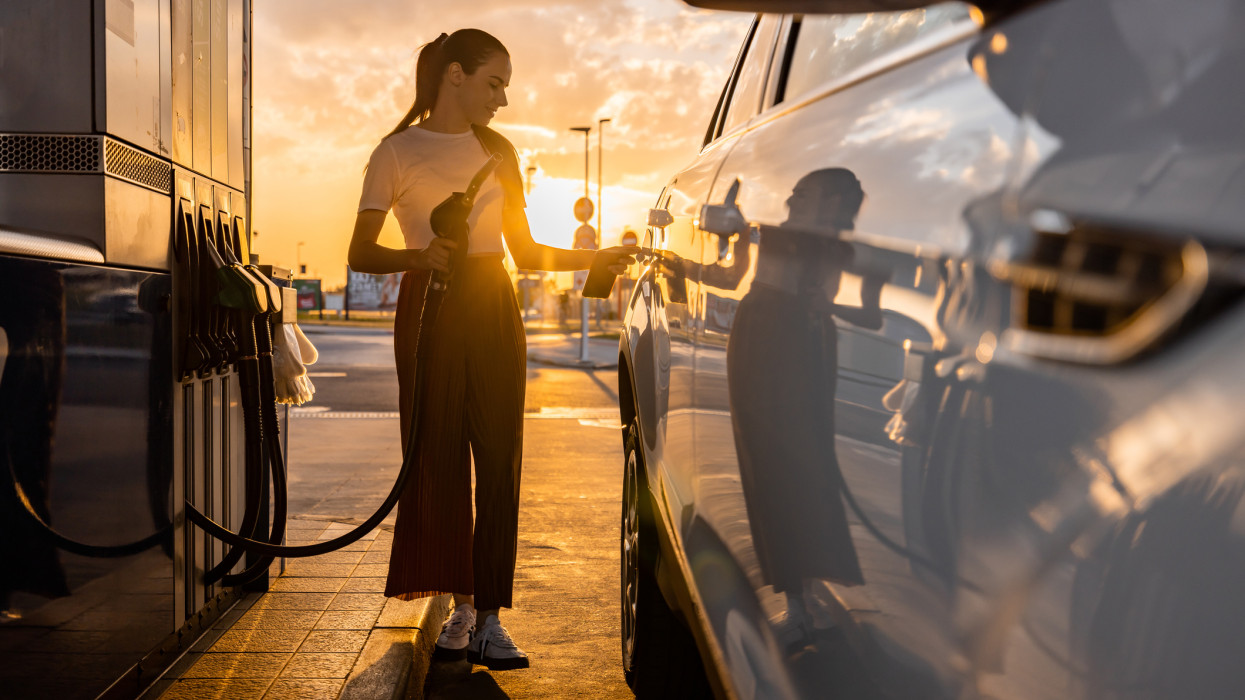 Beautiful young woman refueling gas tank while standing next to car at fuel pump during sunset