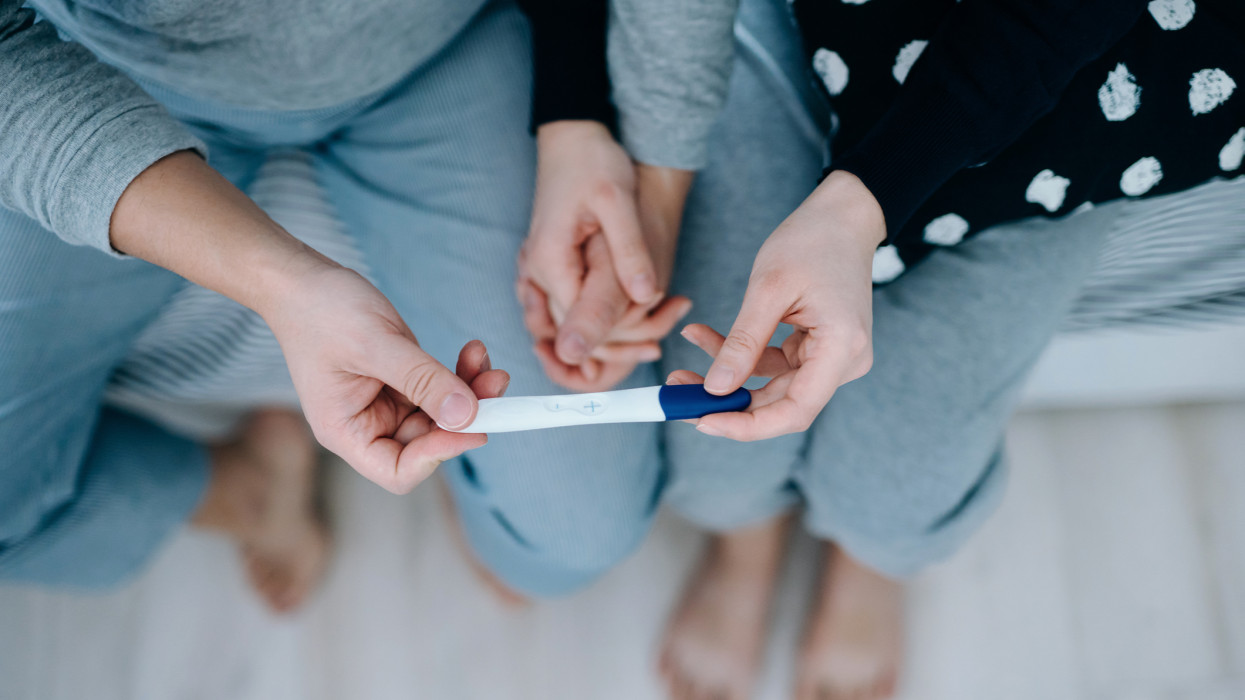 Overhead view of an affectionate young Asian couple sitting on the bed, holding hands and holding a positive pregnancy test together. Its finally happening. The long-awaited news. Life events, fertility and family concept