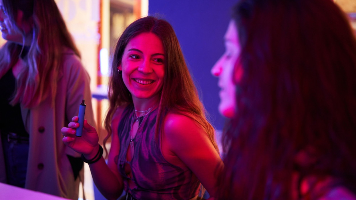Woman sitting at the bar of a discotheque with an electronic cigarette next to friends
