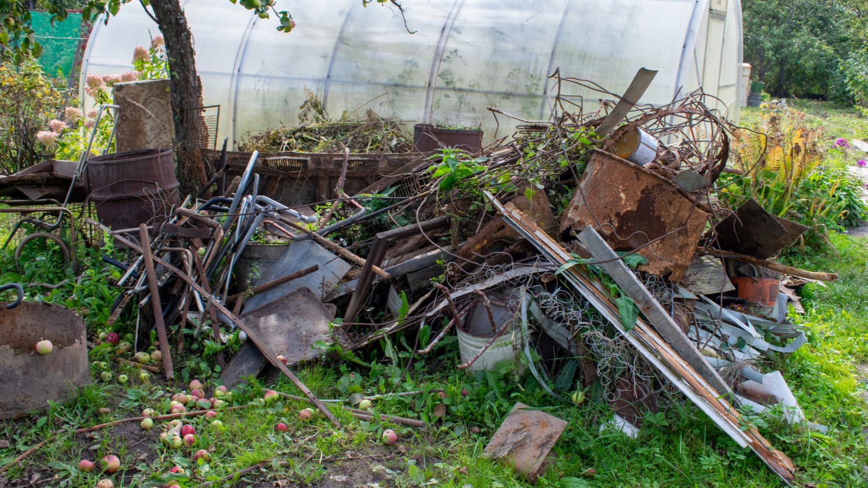 Garbage pile with iron and plastic on the grass. Heap with old rusty iron, tanks, containers, springs. Greenhouse in the country plot. Fresh apples on the grass.