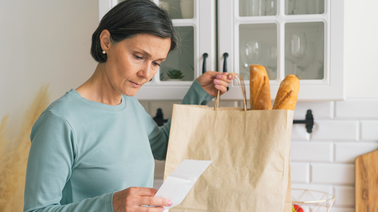 Senior adult female worried while looking at receipt from grocery store. Rise in price of products concept. Buying consumer goods during inflation. Facing economic downturn. Kitchen unit and paper bag on background, horizontal. closeup