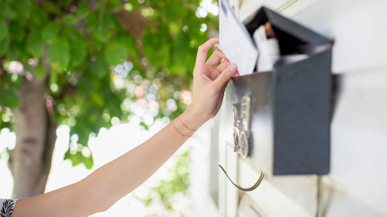 Close up of young womans arm reaching for mail in residential mailbox mounted on exterior wall