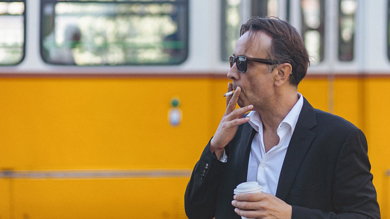 Close-up of a handsome, mature man smoking cigarette and holding disposable cup of coffee while while the yellow tramway passes beside him. The man is pensive while looking away. He is dressed in full black suit and wears black sunglasses. The shot is executed with available natural light, and the copy space has been left.