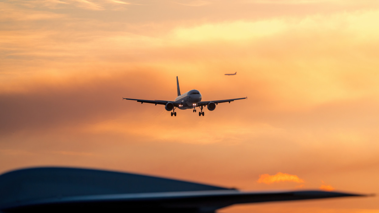 This photograph captures the dynamic nature of an airport during sunset, with planes arriving and departing against a colorful sky. The blurred tail of a parked plane adds movement to the image, while the focused shot of an incoming airplane captures the excitement of air travel.