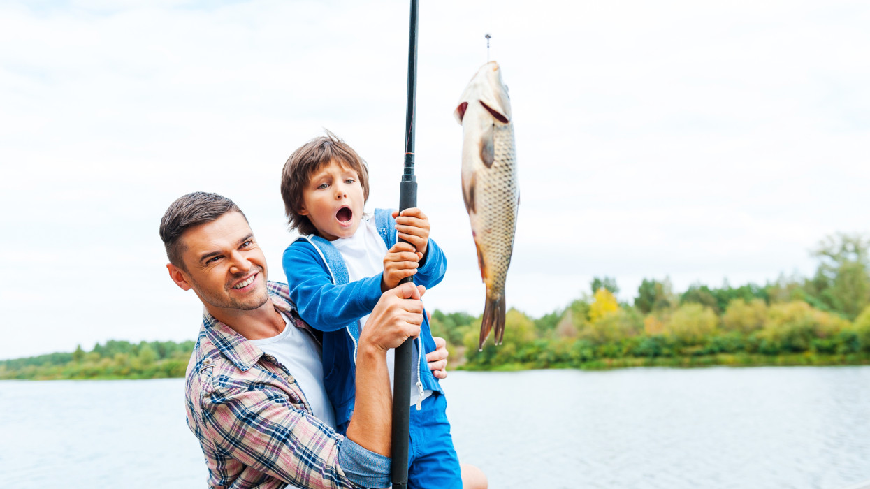 Father and son stretching a fishing rod with fish on the hook while little boy looking excited and keeping mouth open