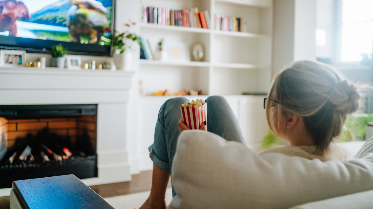 Blond woman with hair bun watching TV on a cozy sofa against fireplace indoors