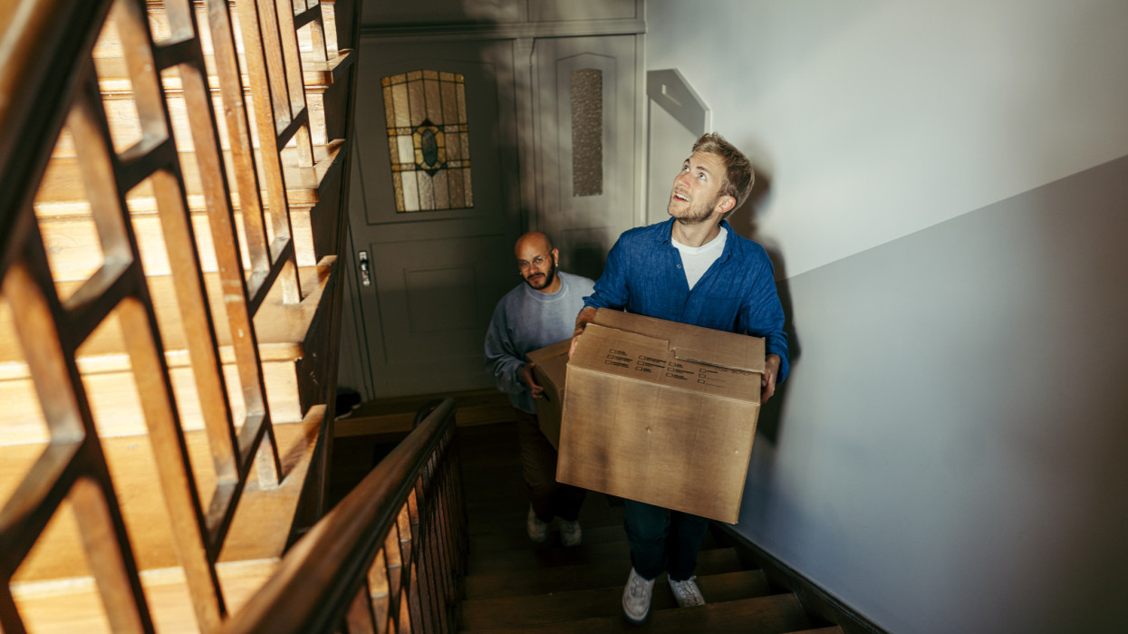 Happy male friends relocating into a new apartment. Two men carrying cardboard boxes and walking up the stairs to their new home.